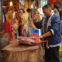 Market butcher preparing meat at a chopping block surrounded by hanging carcasses.